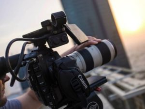 Close-up of a person operating a professional Canon video camera with a large zoom lens, filming an outdoor scene at sunset.