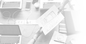 Two people exchange a printed wireframe design over a cluttered desk with laptops, notebooks, and a smartphone, indicating a collaborative web design meeting.