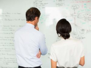Two people standing and analyzing a whiteboard filled with diagrams and notes, viewed from behind.