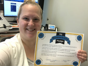 A woman smiling and holding an "Accessibility Pledge" certificate in an office setting. A computer and desk clutter are visible in the background.