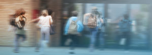 A group of students with backpacks run toward the entrance of a school building. The image has a motion blur effect and a soft blue gradient overlay.