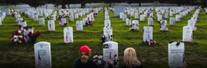 Two people sit in front of decorated headstones at a military cemetery. Rows of white gravestones with American flags and flowers stretch into the distance.
