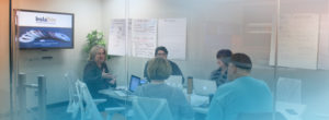 A team of five people sits in a conference room brainstorming for an InstaTrim campaign. Notes are written on large sheets of paper on the walls, and a screen displays the InstaTrim logo and product image.