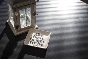 A small decorative lantern and a dish with the words "the best is yet to be" sit on a dark surface with sunlight casting striped shadows.