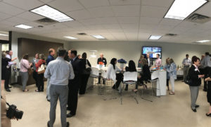 A group of people gathers in a modern office space, engaging in conversation. Some are standing, while others sit at tables. A TV screen is visible in the background.