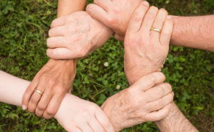 Six hands of different skin tones grasp each other's wrists in a connected circle, symbolizing unity, against a background of grass.