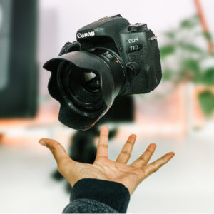 A Canon EOS 77D camera appears to be floating above an outstretched hand, with a blurred background of indoor plants and soft lighting.