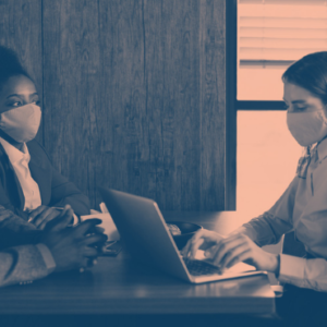 Three people wearing face masks sit at a table during a meeting, with two individuals on one side and one on the other, discussing something over a laptop.