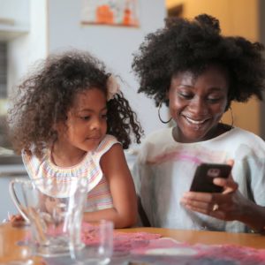 A woman smiles while looking at her phone, sitting next to a young girl who is watching with interest. They are seated at a kitchen table with glasses and a mug.