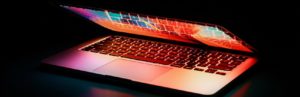 A partially closed laptop in a dark setting emits a colorful glow from its screen and keyboard, casting vibrant reflections on the surface below.