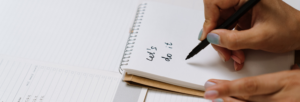 A person writes "Let's do it" in a spiral notebook with a black pen. Lined paper and a to-do list are visible in the background.