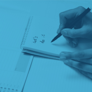 A person writes "Let's do it" in a spiral notebook with a black pen. Lined paper and a to-do list are visible in the background.