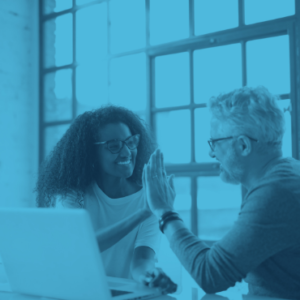 Two colleagues, one woman and one man, smiling and giving each other a high five while sitting in front of a laptop in a bright office space.