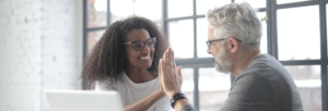 Two colleagues, one woman and one man, smiling and giving each other a high five while sitting in front of a laptop in a bright office space.
