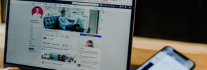 A person browsing a Facebook profile on a laptop, with a smartphone visible in the foreground. The profile features a cover photo of a man sitting on a couch.