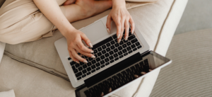 A person sitting cross-legged on a couch types on a laptop placed on their lap, with their hands resting on the keyboard. The setting is casual and relaxed.