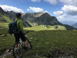 A cyclist in a helmet and green jacket pauses on a mountain trail, looking out at grassy slopes and rugged peaks under a partly cloudy sky.