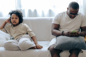 a child and a man sitting on a couch, the man is looking at his phone