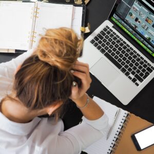 Woman sitting at her laptop, her head in her hands. Frustrated. She has eyeglasses and two notebooks on the desk near her.