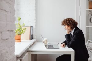 A woman sits at a desk, starting down at her laptop. He hand is in her hair, implying she may be frustrated or focusing. The room is white, a window across from her white desk, with a plant in the window.