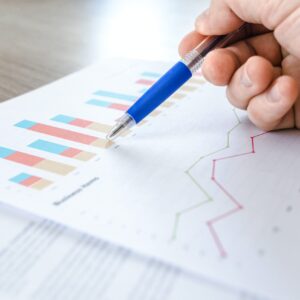 A hand holds a pen, touching a page of colorful graphs and analytics.