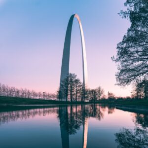 The Gateway Arch in St. Louis, Missouri, set against a colorful sunset sky with pink and orange clouds. The city skyline is visible beneath the arch, with buildings and waterfront lights reflecting on the water in the foreground.