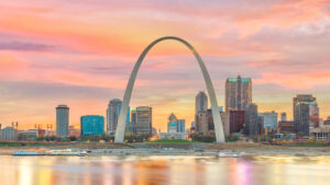 The Gateway Arch in St. Louis, Missouri, set against a colorful sunset sky with pink and orange clouds. The city skyline is visible beneath the arch, with buildings and waterfront lights reflecting on the water in the foreground.