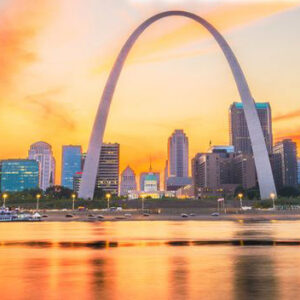 The Gateway Arch in St. Louis, Missouri, set against a colorful sunset sky with pink and orange clouds. The city skyline is visible beneath the arch, with buildings and waterfront lights reflecting on the water in the foreground.