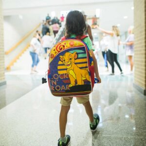 A young child with a "Lion King" backpack featuring Simba and the text "Rock 'N' Roar" walks down a school hallway, with people in the background.